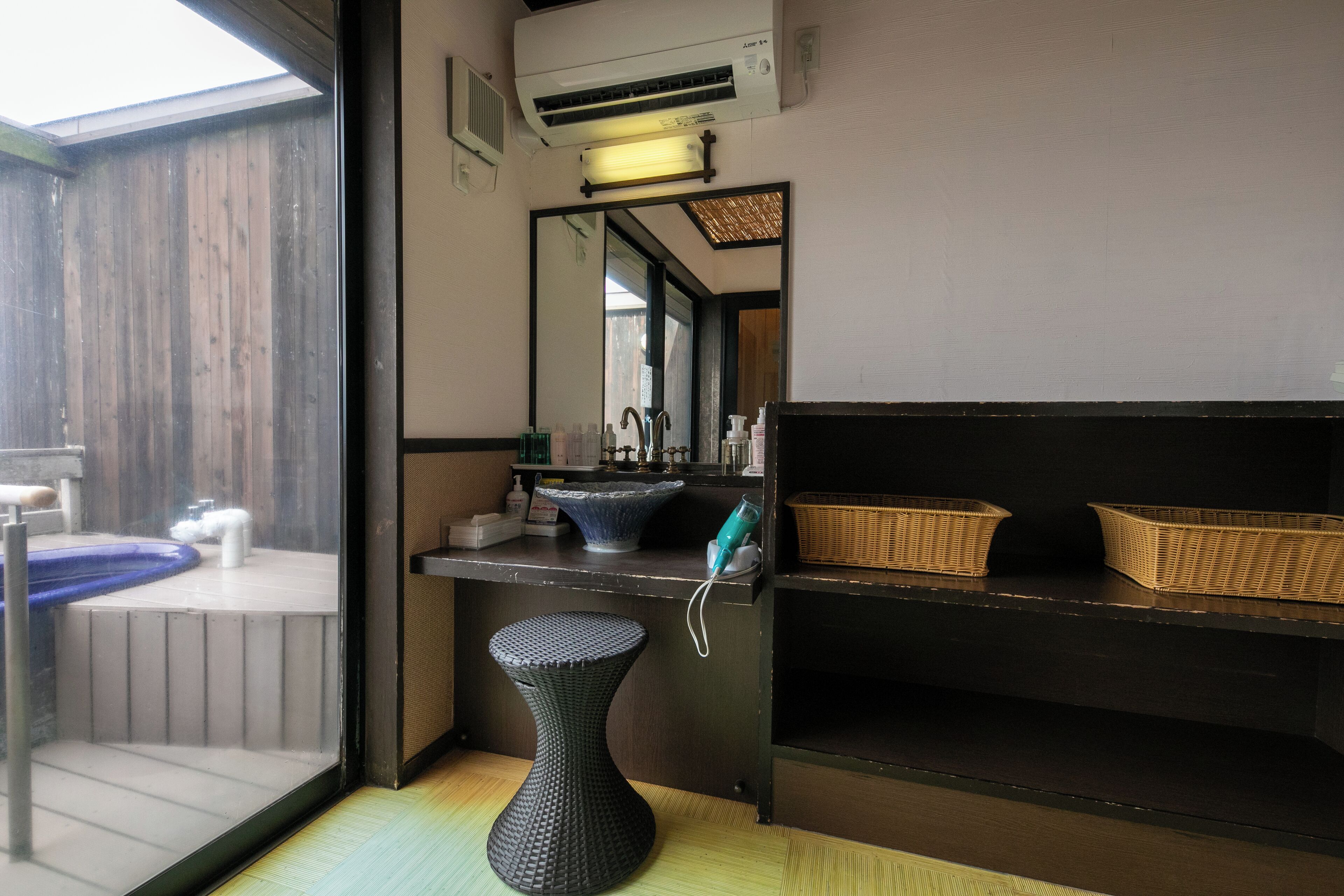 Un baño con un espejo grande, un lavabo, una bañera azul, un taburete negro y cestas de mimbre en estantes.