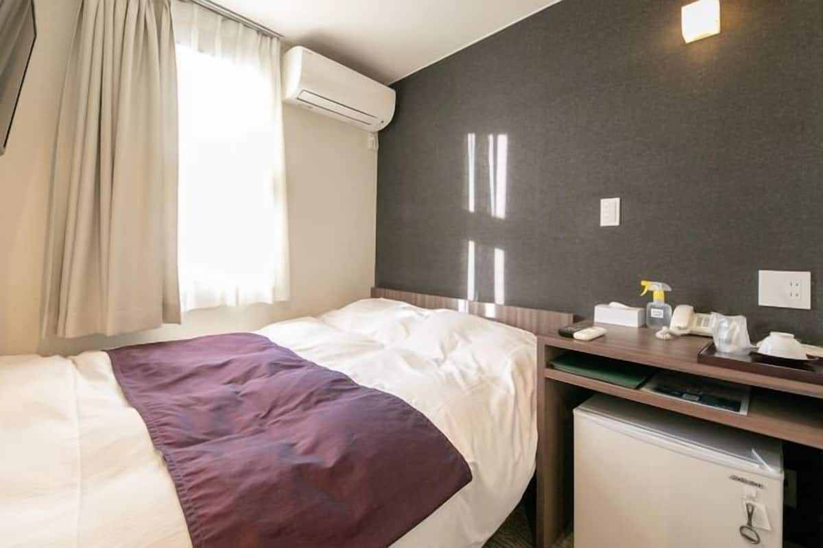 A hotel room with a bed, a desk with a chair, a small refrigerator, an air conditioning unit, and a window with curtains.