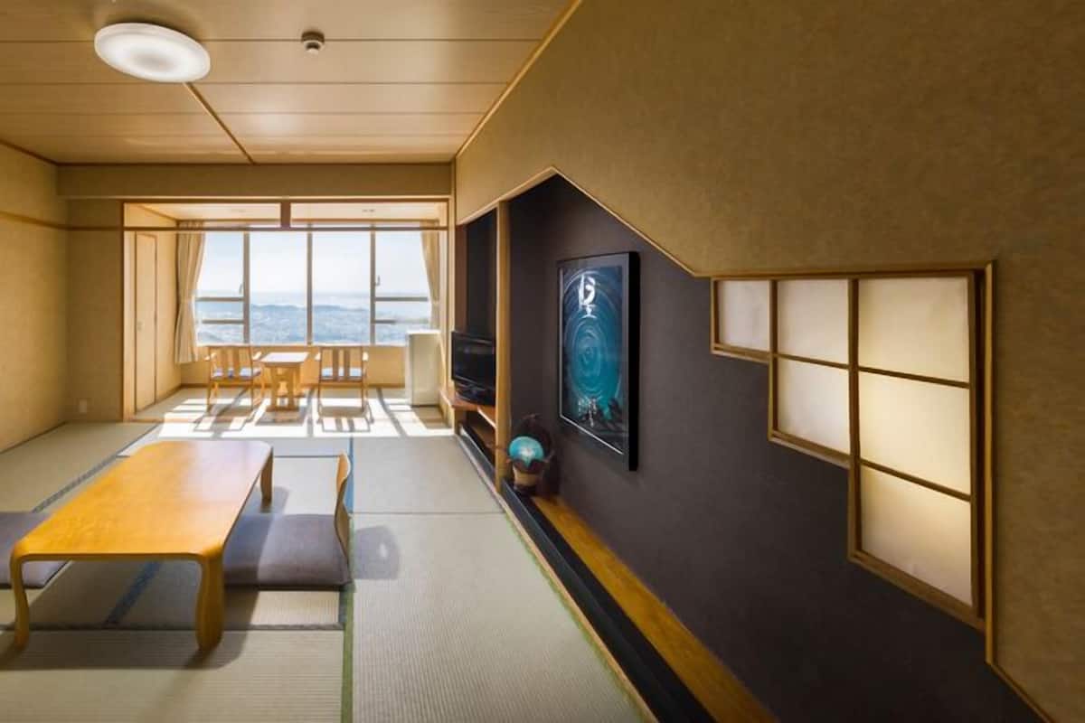 A traditional Japanese room with tatami flooring, a low table, chairs, a television, and a large window offering a view.