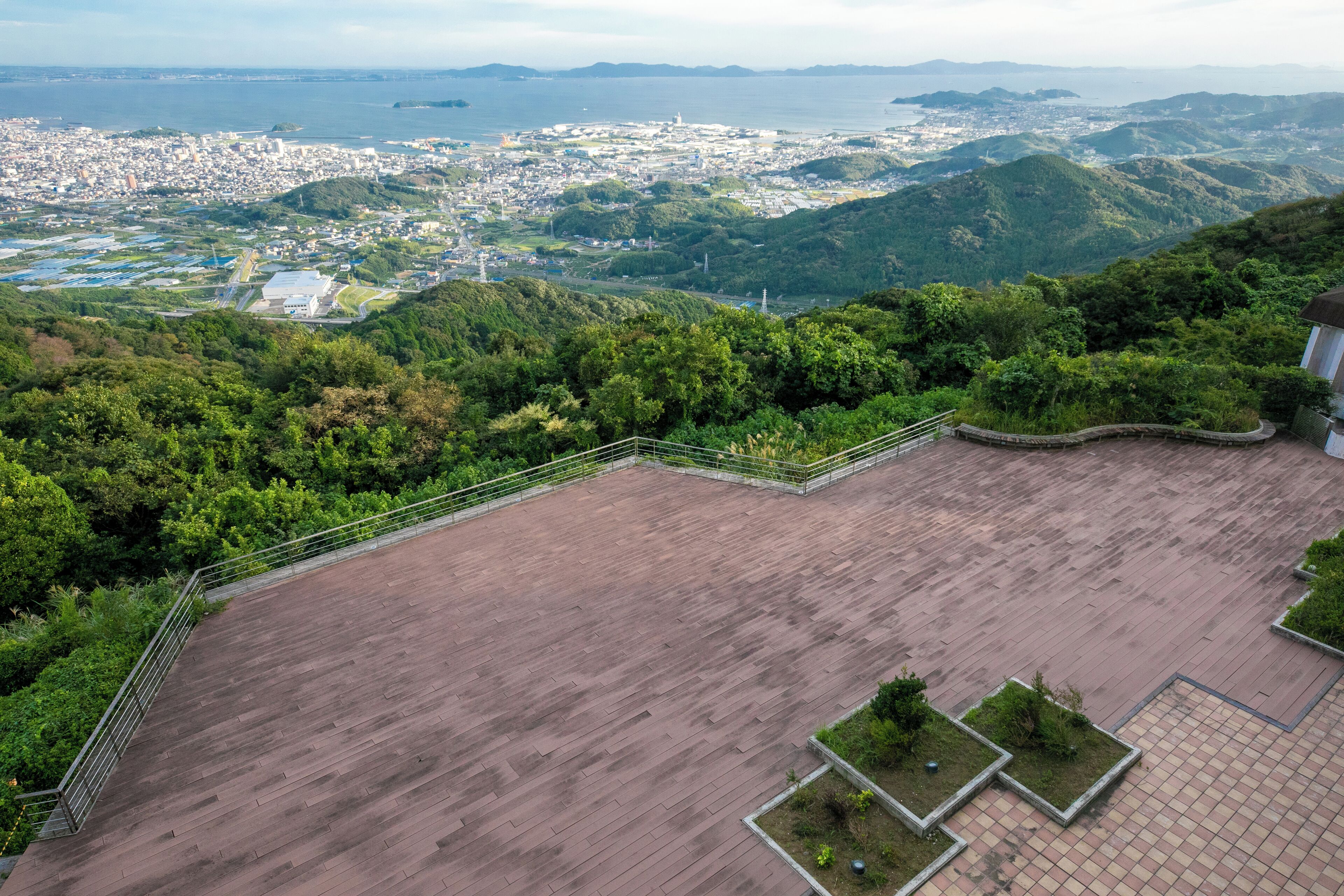 A panoramic view of a city from a high vantage point, featuring a large open plaza with a metal railing, surrounded by lush greenery and hills.