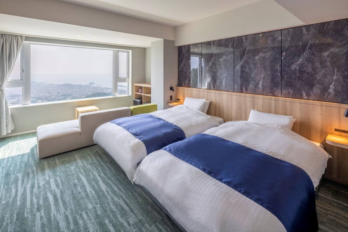 A hotel room with two beds, a large window, a marble wall, and a city view.