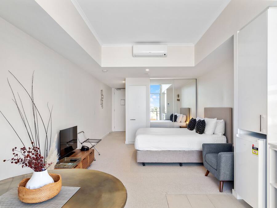 A modern hotel room with a bed, a sofa, a dining table, a TV, and a kitchen area.