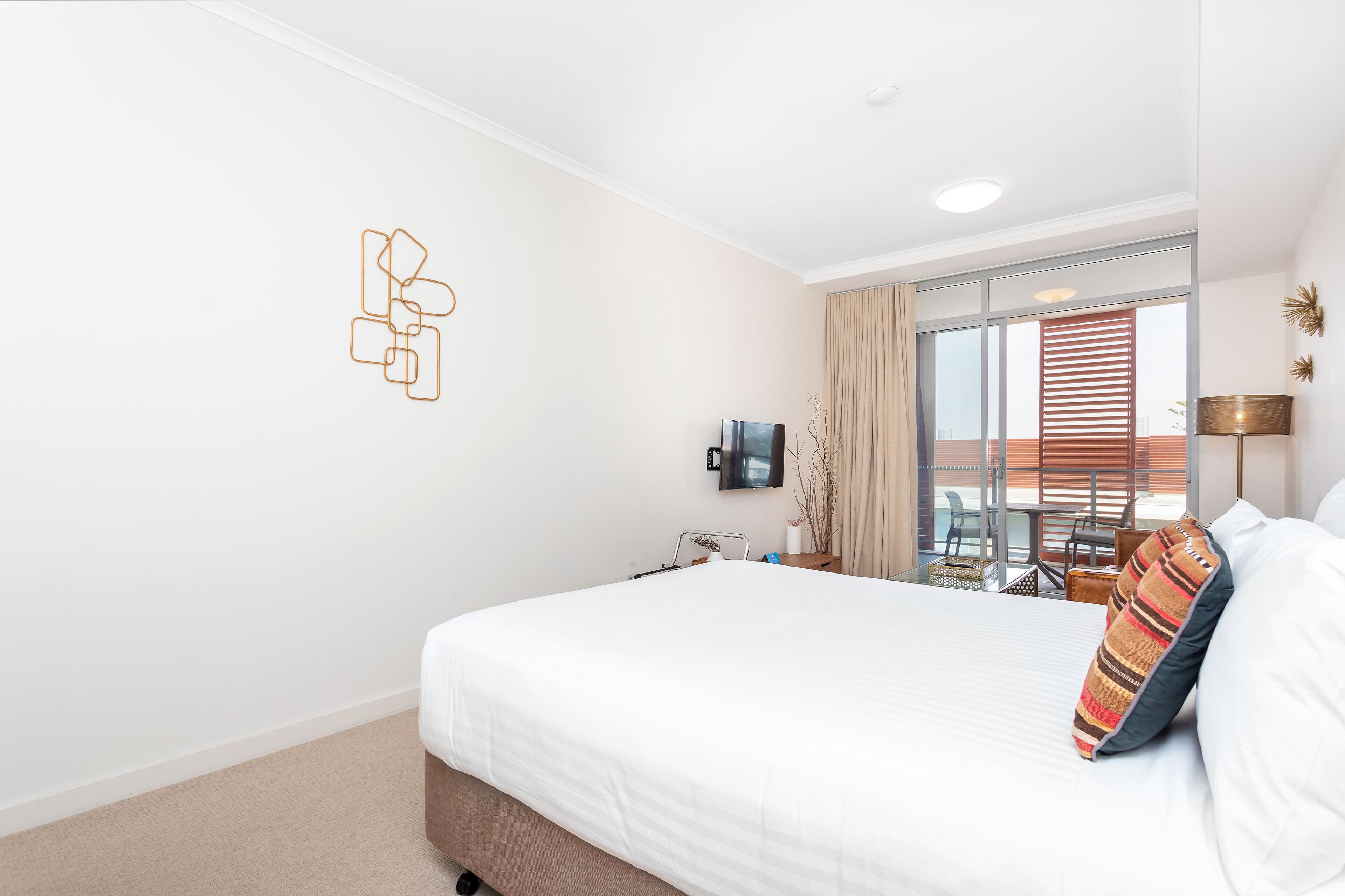 A hotel room with a large bed, a TV mounted on the wall, a balcony with a table and chairs, and a window with curtains.