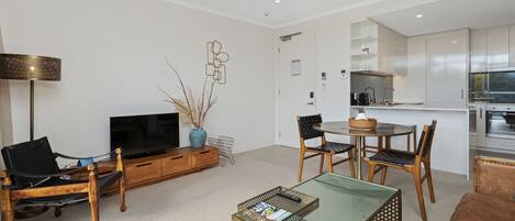 A modern living room with a flat-screen TV, wooden furniture, a glass coffee table, and a kitchen area with white cabinets and a stainless steel oven.