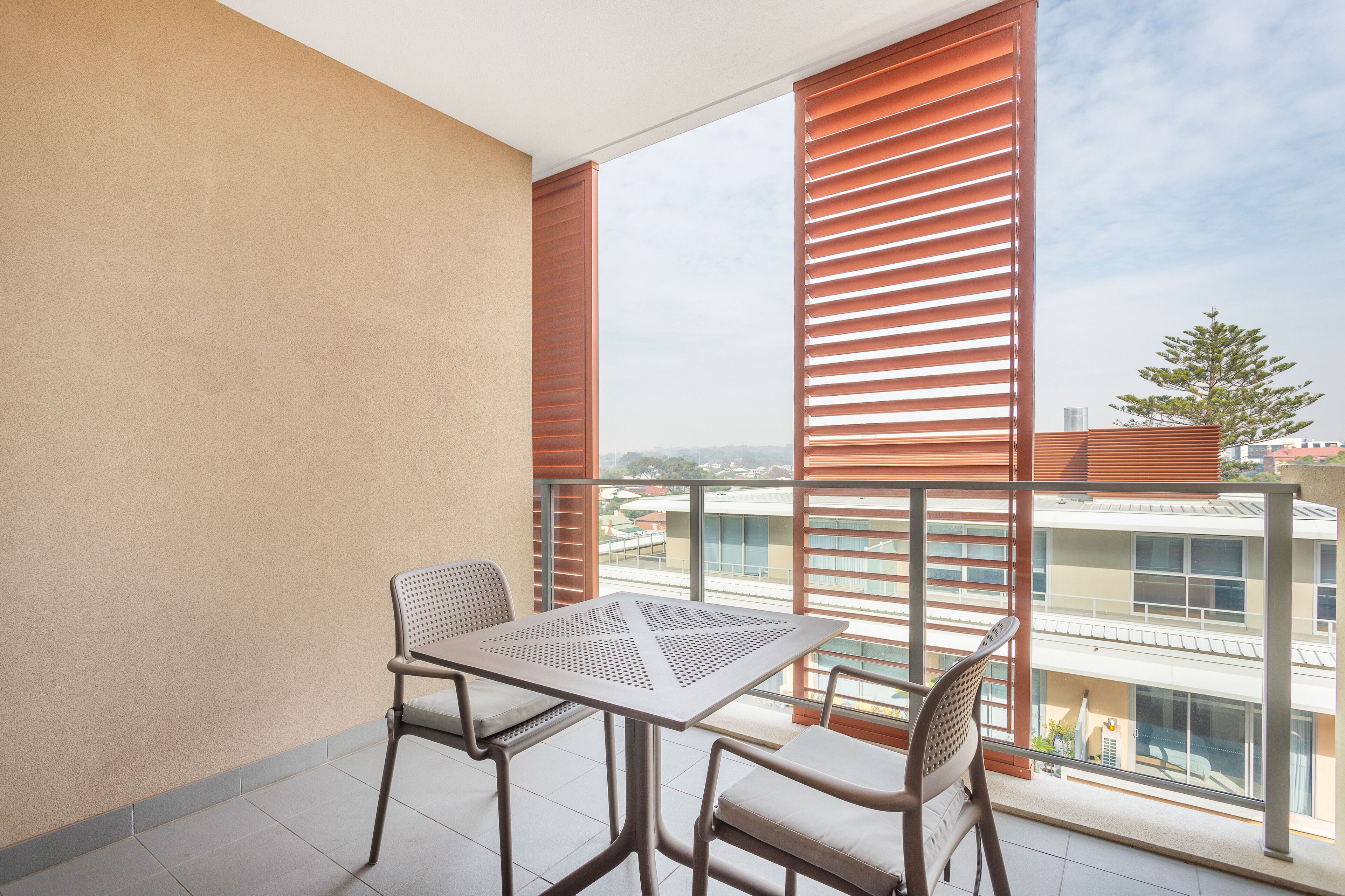 Uma varanda com mesa e cadeiras, janelas amplas com persianas e vista para edifícios.