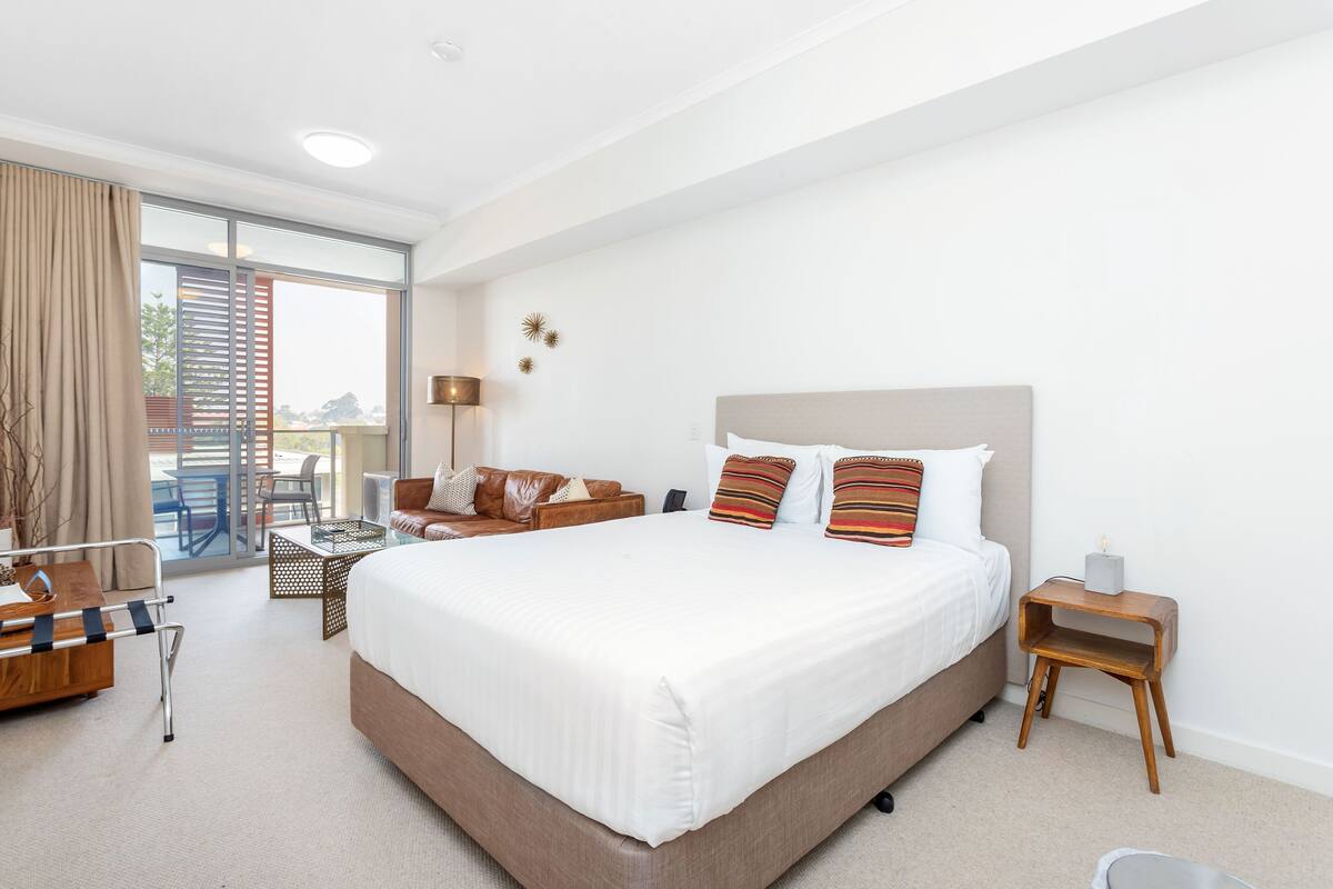 A modern hotel room with a large bed, a sofa, a small table, and a chair. There is a balcony with a view outside.