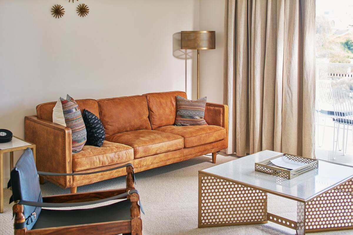 A living room with a brown leather sofa, patterned cushions, a wooden rocking chair, a glass coffee table, and a floor lamp.