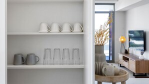 A modern interior with white shelves displaying glassware and mugs, a wooden sideboard with decorative items, and a view through an open door. - Nautica Residences Fremantle (East Fremantle)