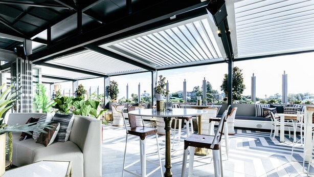 A modern outdoor lounge area with a sofa, dining table, and chairs, surrounded by potted plants and offering a view of the city skyline. - Nautica Residences Fremantle (East Fremantle)