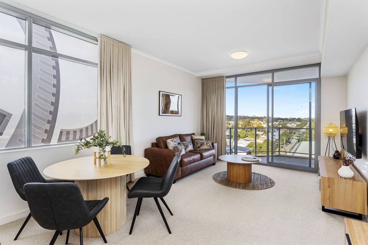 A modern hotel room with a large window, a brown leather sofa, a dining table with chairs, and a TV on a wooden stand.
