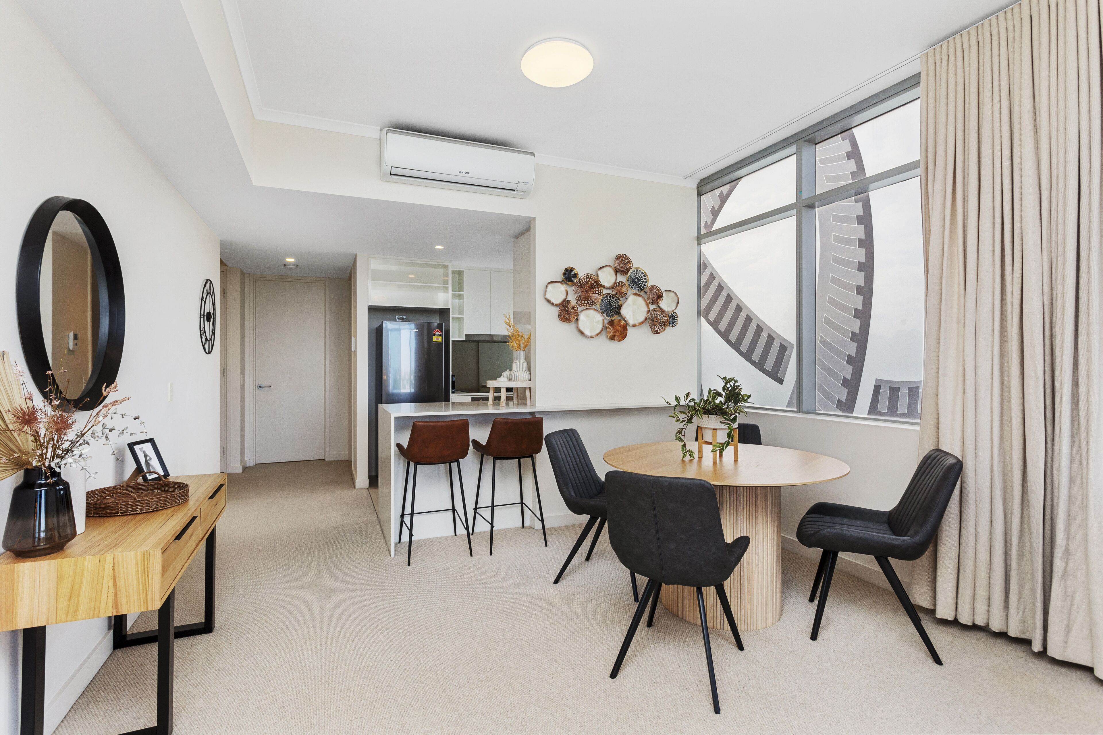A modern living room with a dining area, a large window with curtains, a kitchen in the background, and a ceiling-mounted air conditioner.
