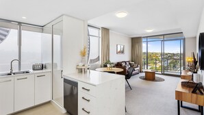 A modern kitchen with a built-in dishwasher, a dining area with a round table, and a living room with a sofa and a TV. - Nautica Residences Fremantle (East Fremantle)