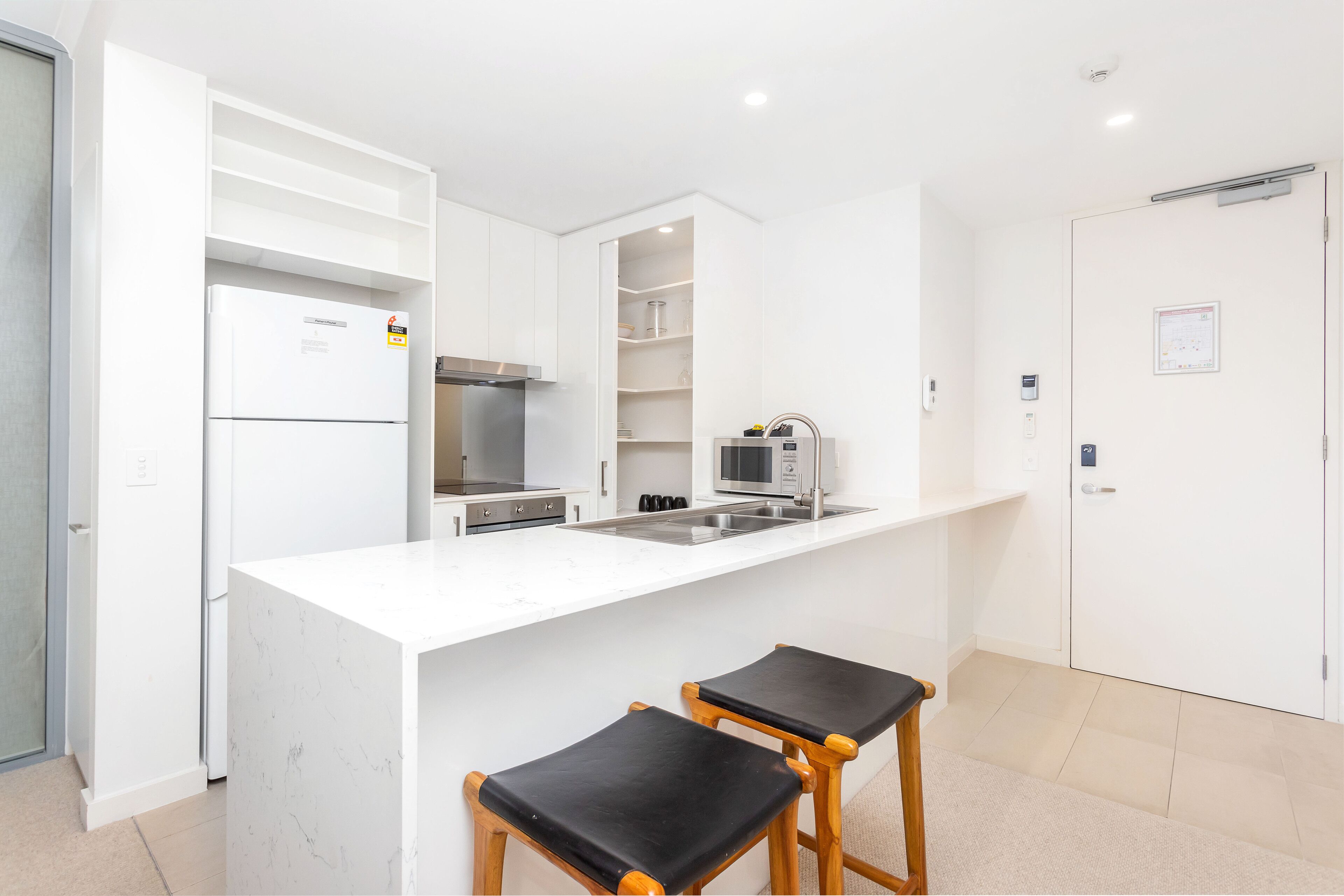Una cocina moderna con muebles blancos, un microondas empotrado y un lavamanos de acero inoxidable. Hay dos taburetes de madera con cojines negros.