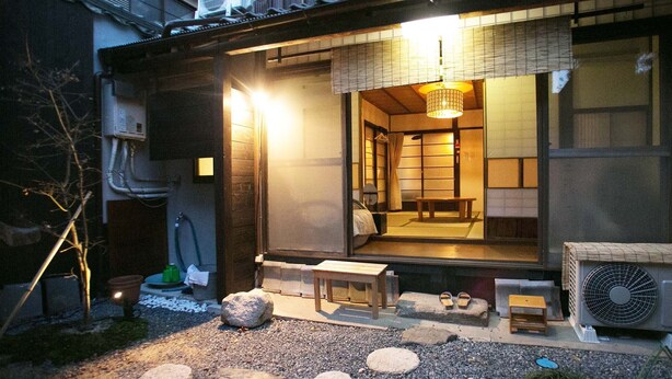 A traditional Japanese ryokan with tatami mat flooring, a wooden exterior, and a sliding door leading to a room with a futon bed. - Guesthouse Hana Nishijin (Kyoto)