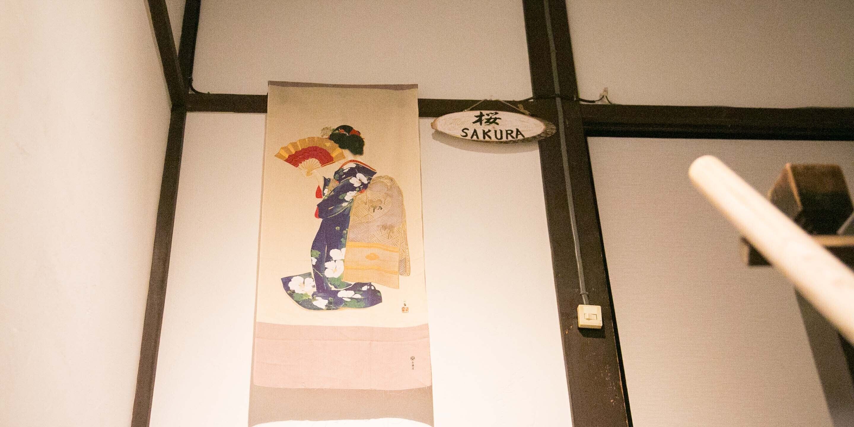 A wall with a hanging artwork depicting a figure in traditional attire, a sign reading "SAKURA," and a white pole.