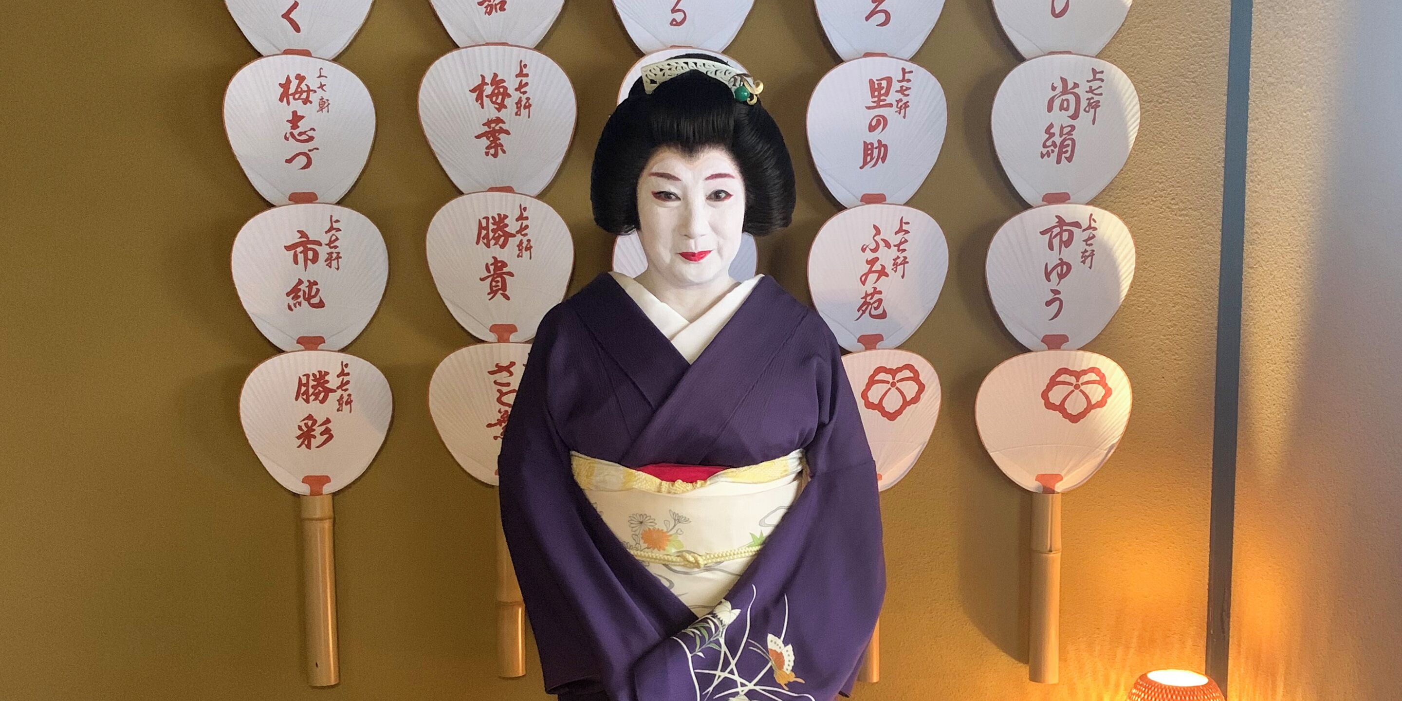 Une personne vêtue d’un costume traditionnel, debout devant un mur décoré d’éventails en papier.