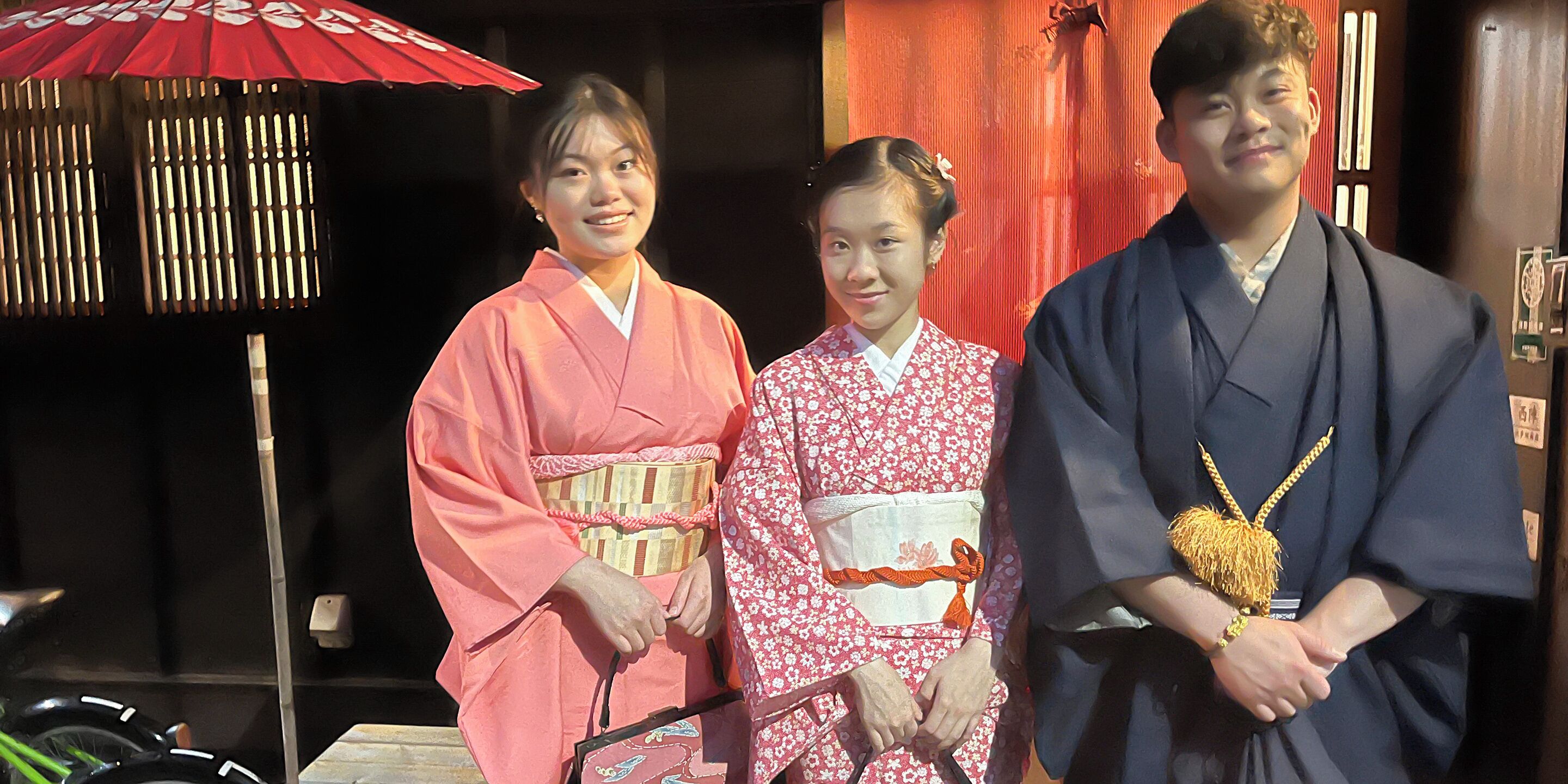 Three individuals in traditional attire standing in front of a building with a red door and a red umbrella.