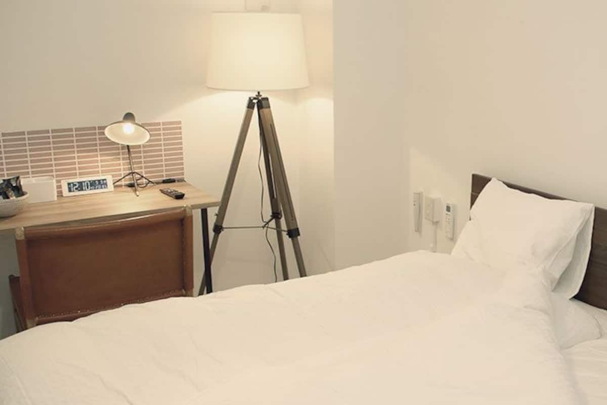 A hotel room with a bed, a bedside table with a lamp, a clock, and a power outlet.