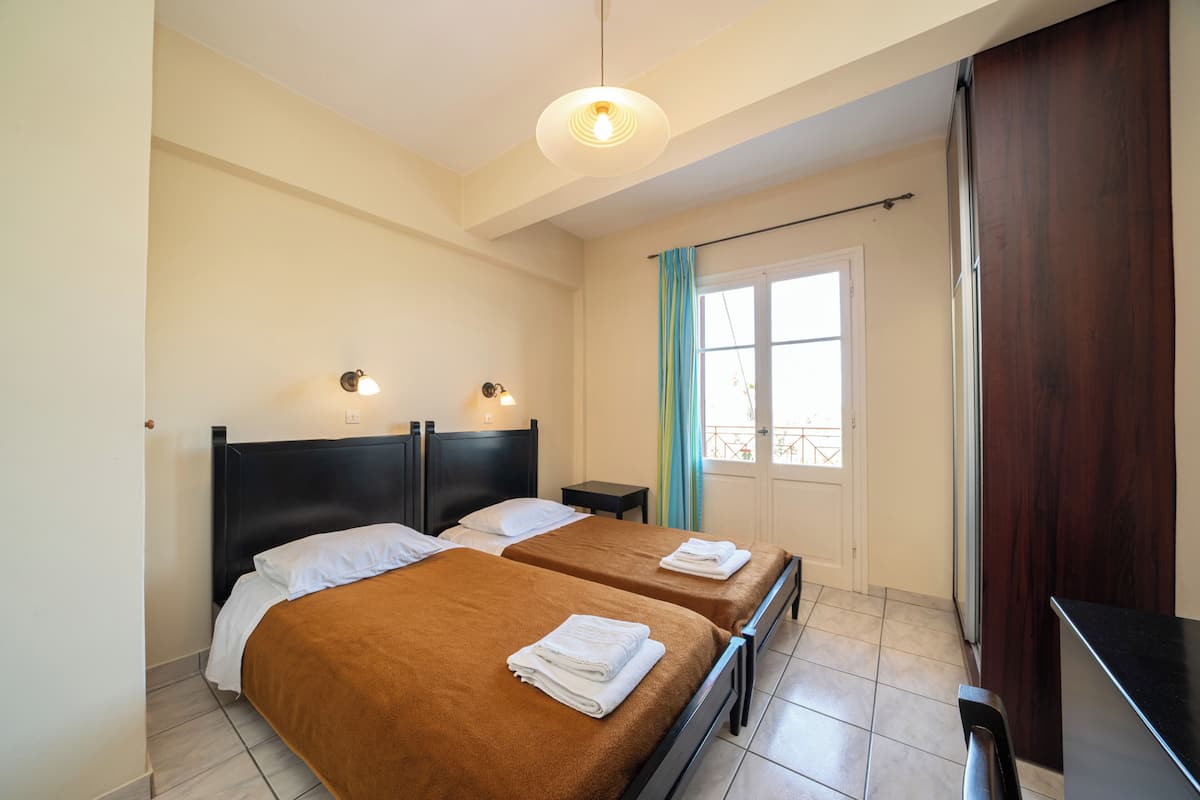 A hotel room with two beds, a small bedside table, a nightstand, a window with curtains, and a door.