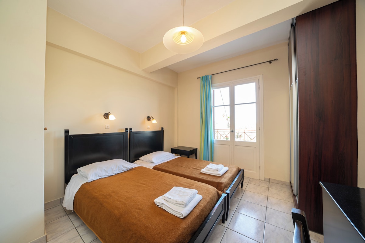 A hotel room with two beds, a small bedside table, a nightstand, a window with curtains, and a door.