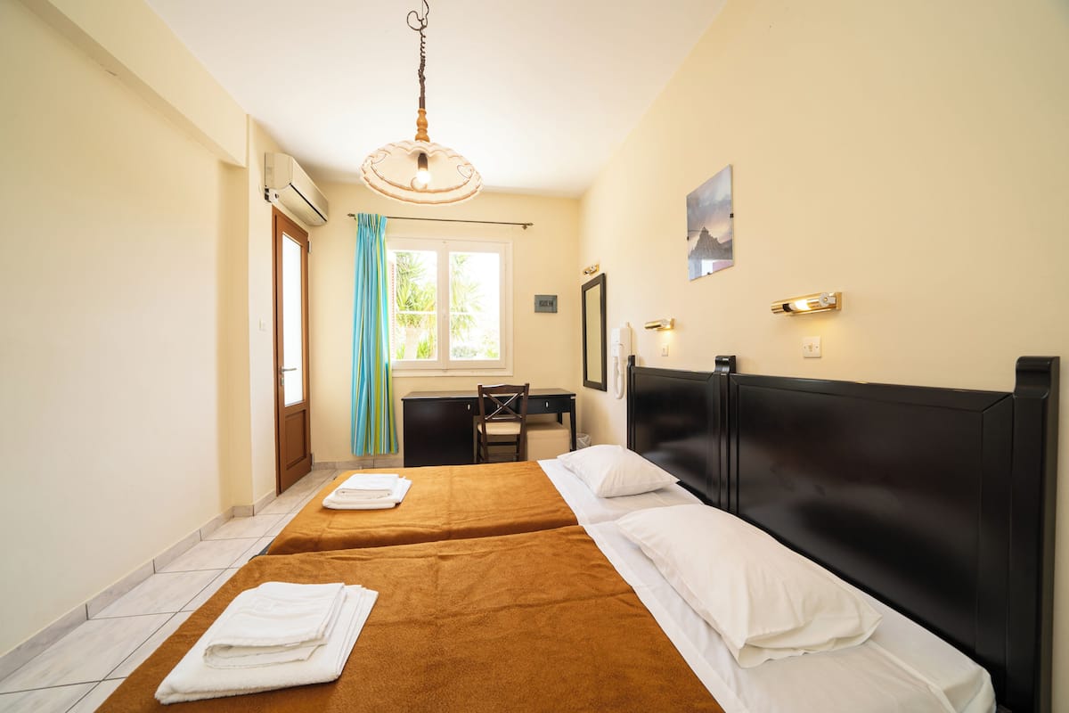 A hotel room with two beds, a desk, a chair, a window with curtains, an air conditioning unit, and a wall-mounted light fixture.