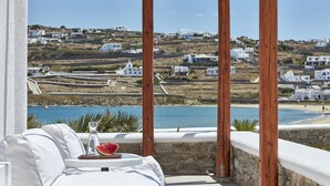 Ein Balkon mit einer weißen Couch, einem Glas Wasser und einer Wassermelone. Blick auf eine Küstenlandschaft mit weißen Gebäuden und blauem Himmel.