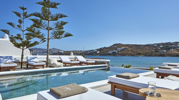 Ein Poolbereich mit Liegestühlen, einem kleinen Baum und Blick auf Meer und Hügel.
