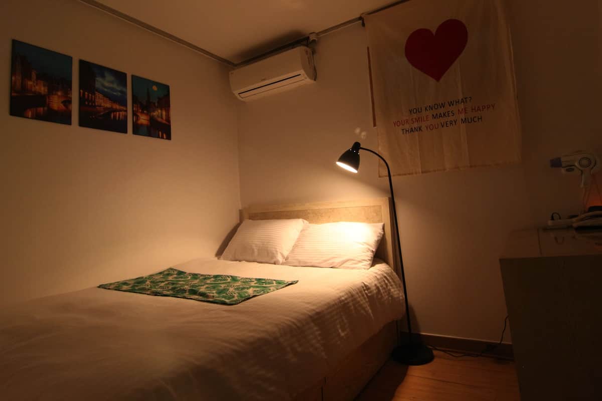 Un dormitorio con cama, mesita de noche, lámpara, aire acondicionado instalado en la pared y un cartel con un corazón y texto.