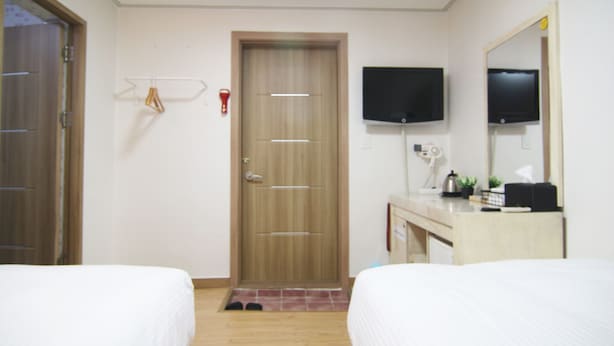 A hotel room with two beds, a wooden door, a television mounted on the wall, and a small desk with a mirror and a plant. - Hotel FA (Seoul)