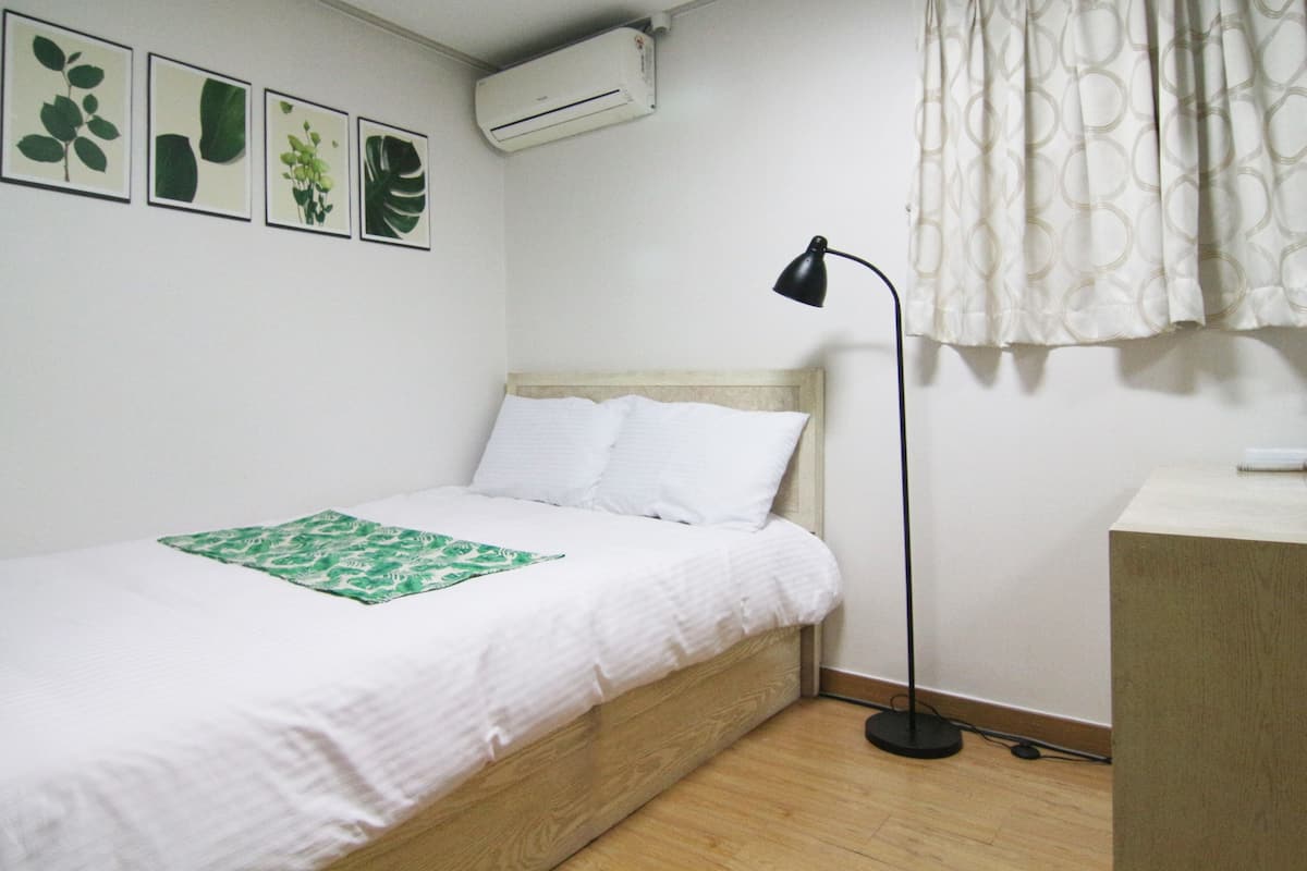Una cama bien hecha con ropa de cama blanca y una manta verde estampada, una lámpara de pie negra y un aire acondicionado instalado en la pared.