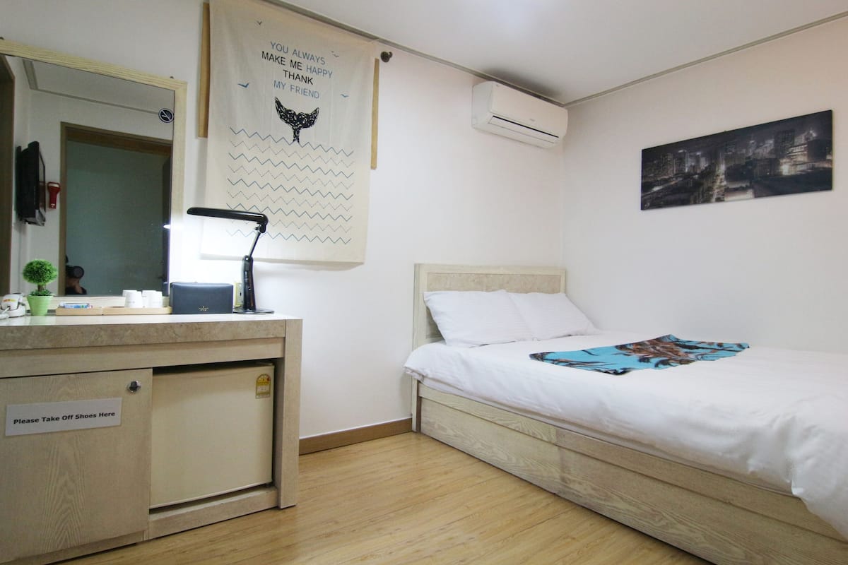 Un dormitorio con una cama, un escritorio con lámpara, un espejo, aire acondicionado y un cuadro de un paisaje urbano.