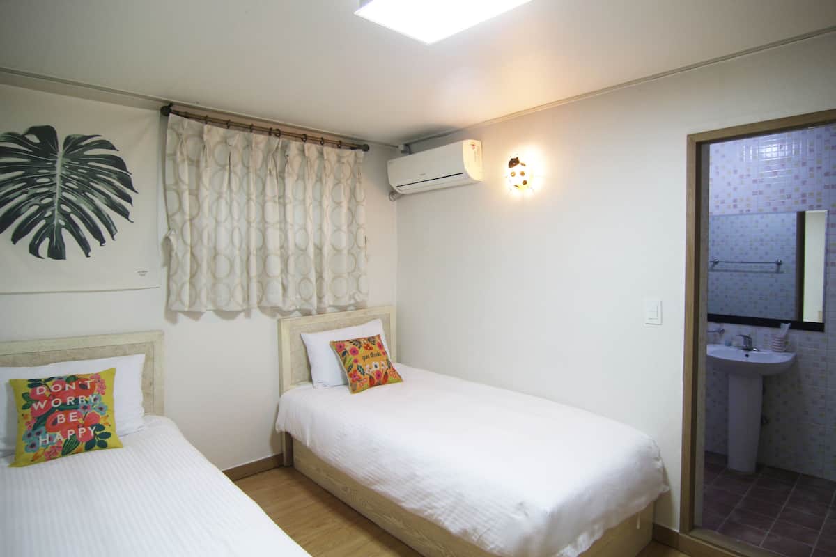 Un dormitorio con dos camas, una planta grande, un equipo de aire acondicionado y un baño visible a través de una puerta abierta.
