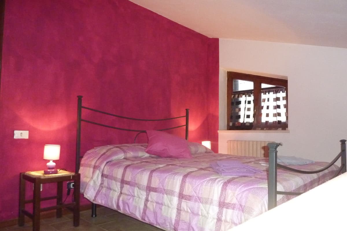 Ein Schlafzimmer mit Bett, Nachttisch, Lampe und einem Fenster mit Blick auf ein Gebäude.
