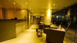 A modern lobby with a reception desk, a sofa, a small table with magazines, and a glass wall with a view of the outside. - Patt Serviced Apartments (Chonburi)