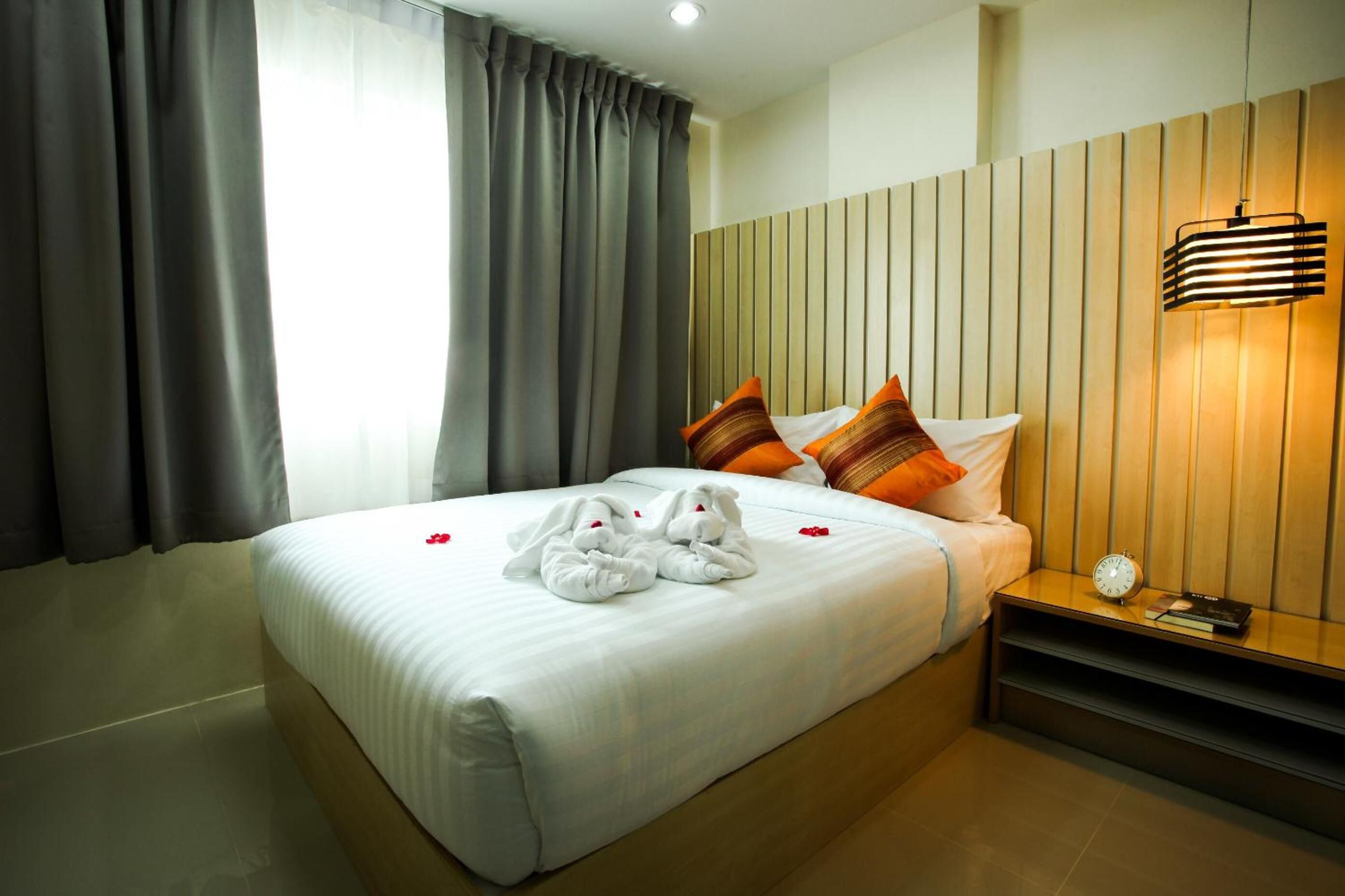 A neatly made bed with white linens and two pillows, a bedside table with a lamp, and a window with curtains.