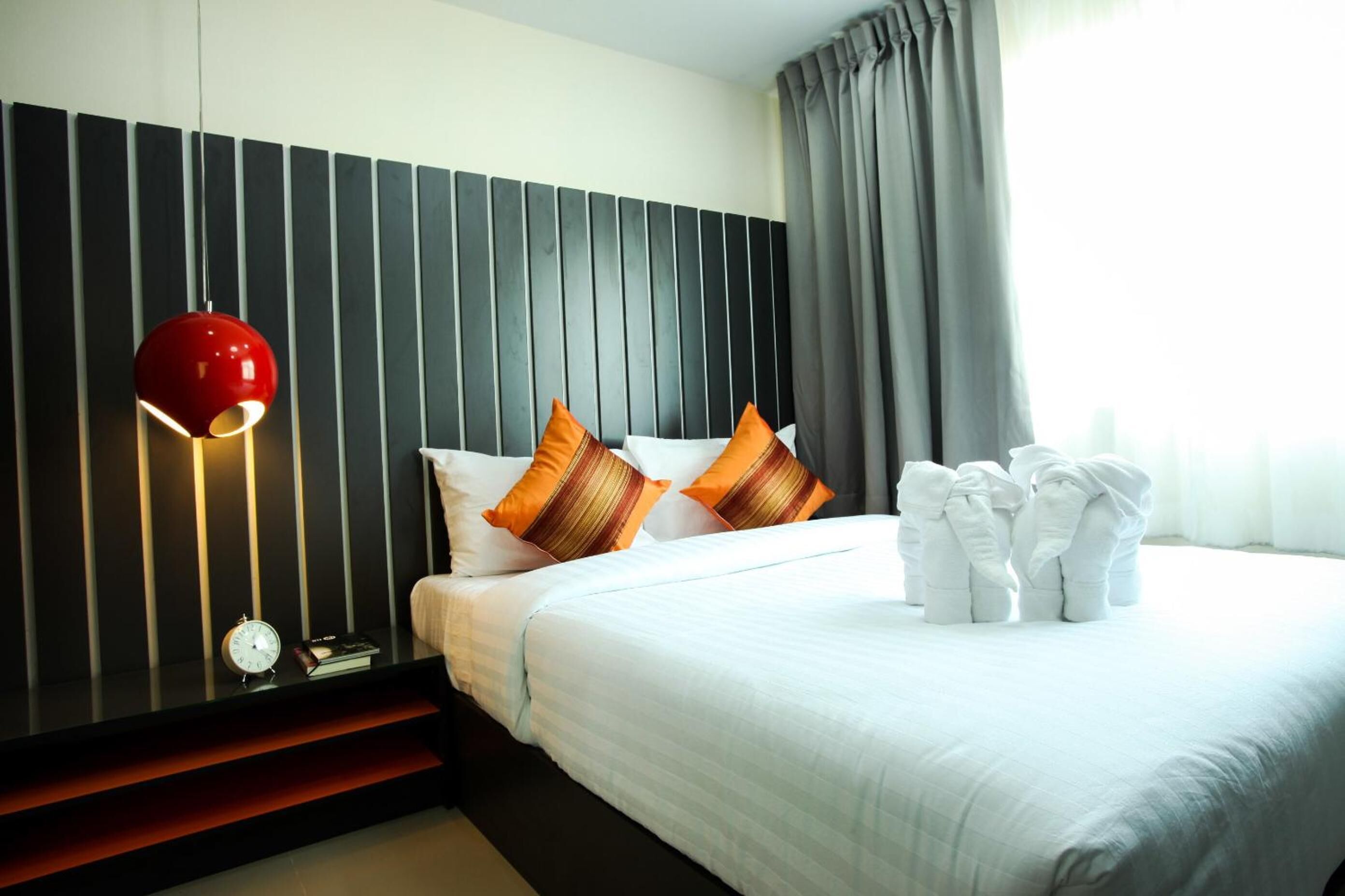 A hotel room with a bed, pillows, a red lamp, a clock, and towels.