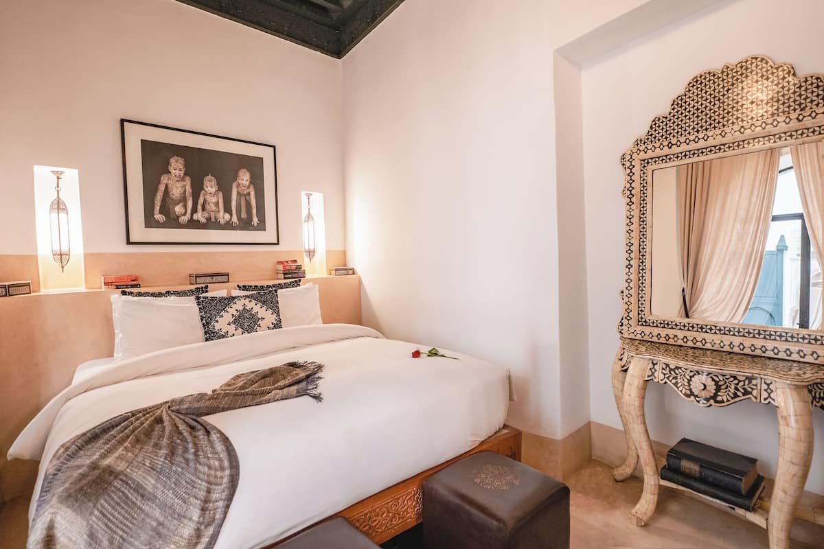 A neatly made bed with a rose, a large ornate mirror, and a framed picture on the wall.
