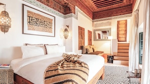 Een ruime slaapkamer met een groot bed, een houten hoofdbord, een geborduurde plaid en een geruit vloerkleed.