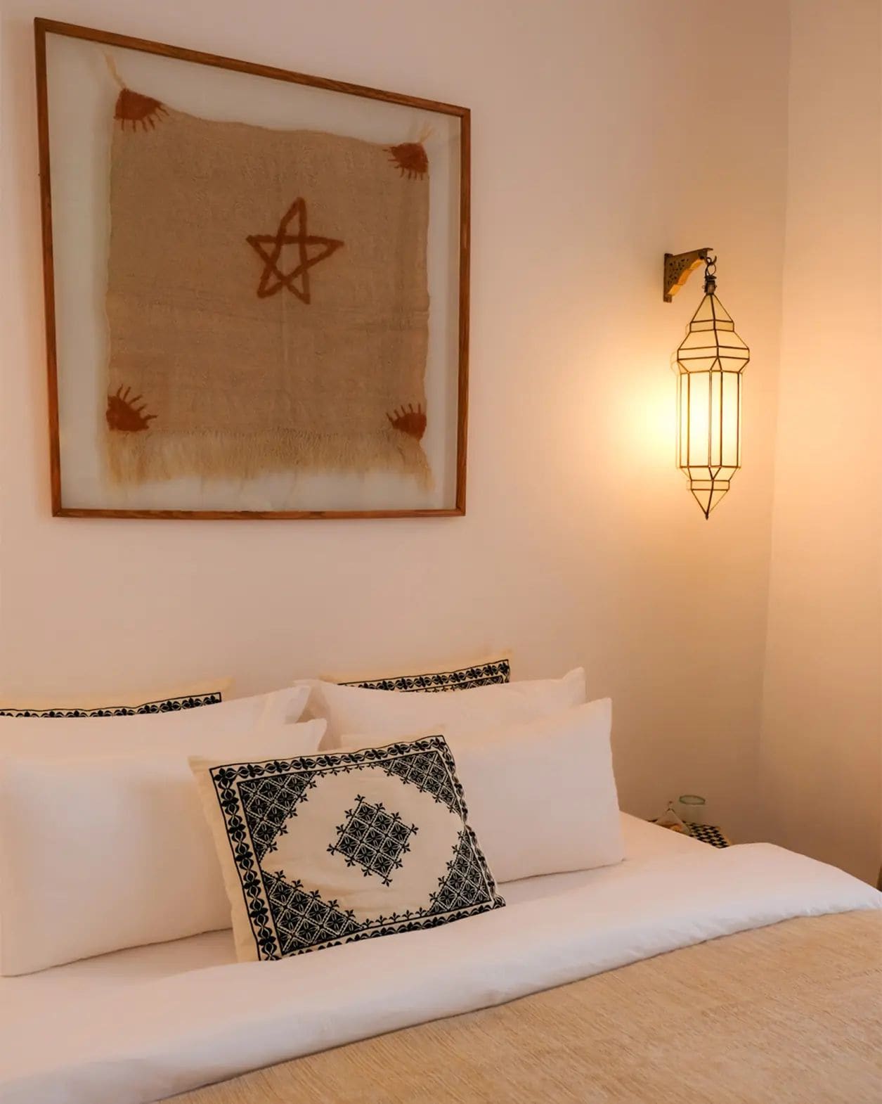 A neatly made bed with white linens and decorative pillows, a framed artwork with a star design on the wall, and a wall-mounted light fixture.