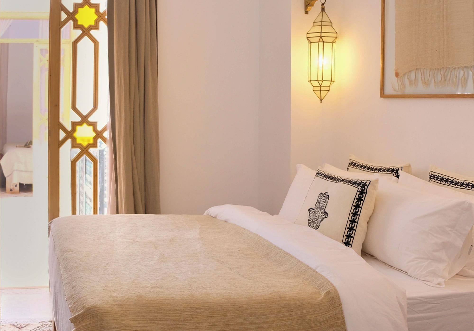 Una cama bien hecha con cubrecama y almohadas color beige, un aplique de pared y una ventana con diseño geométrico.