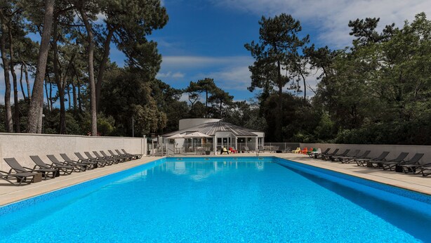 A rectangular swimming pool with lounge chairs on both sides, surrounded by trees and a building with a sloped roof in the background.