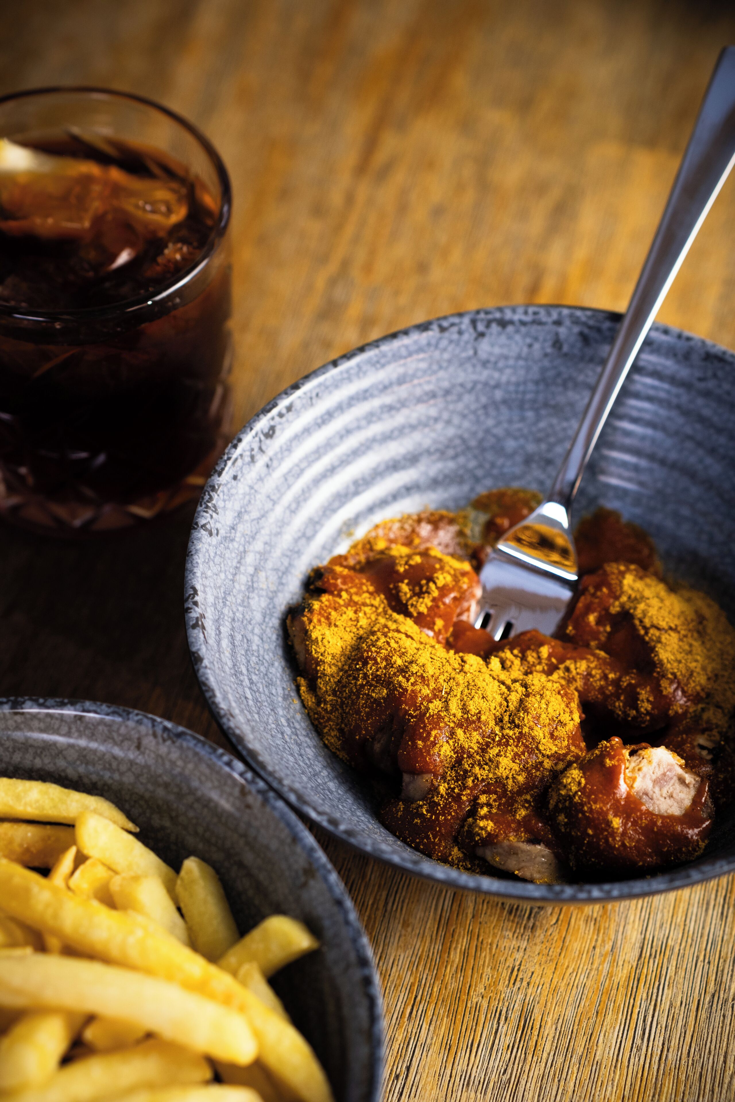 Un tazón de pollo sazonado, un tenedor, un vaso de bebida oscura y un tazón de papas fritas sobre una mesa de madera.