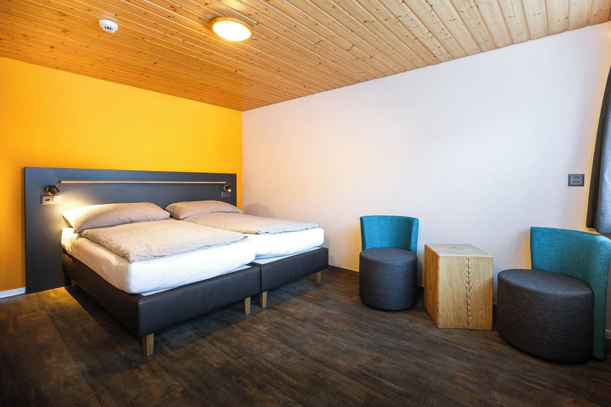 A modern bedroom with a wooden ceiling, a bed with a headboard, two ottomans, and a small wooden side table.