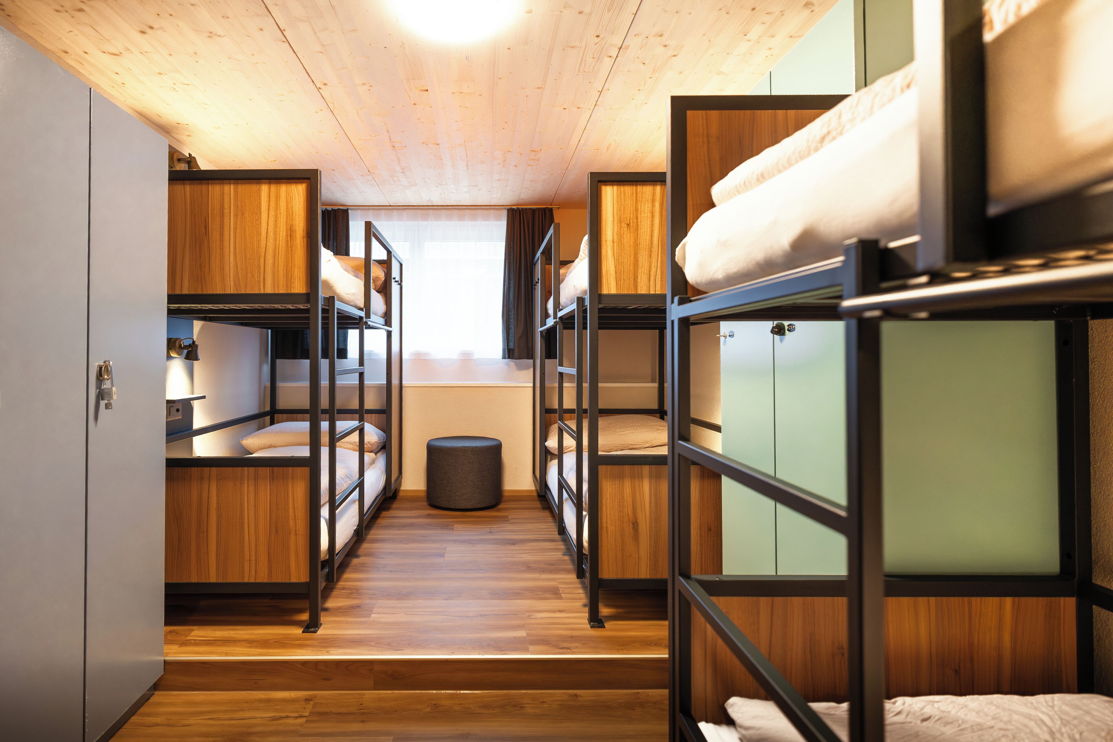 A dormitory room with bunk beds, wooden floors, and a small round stool.