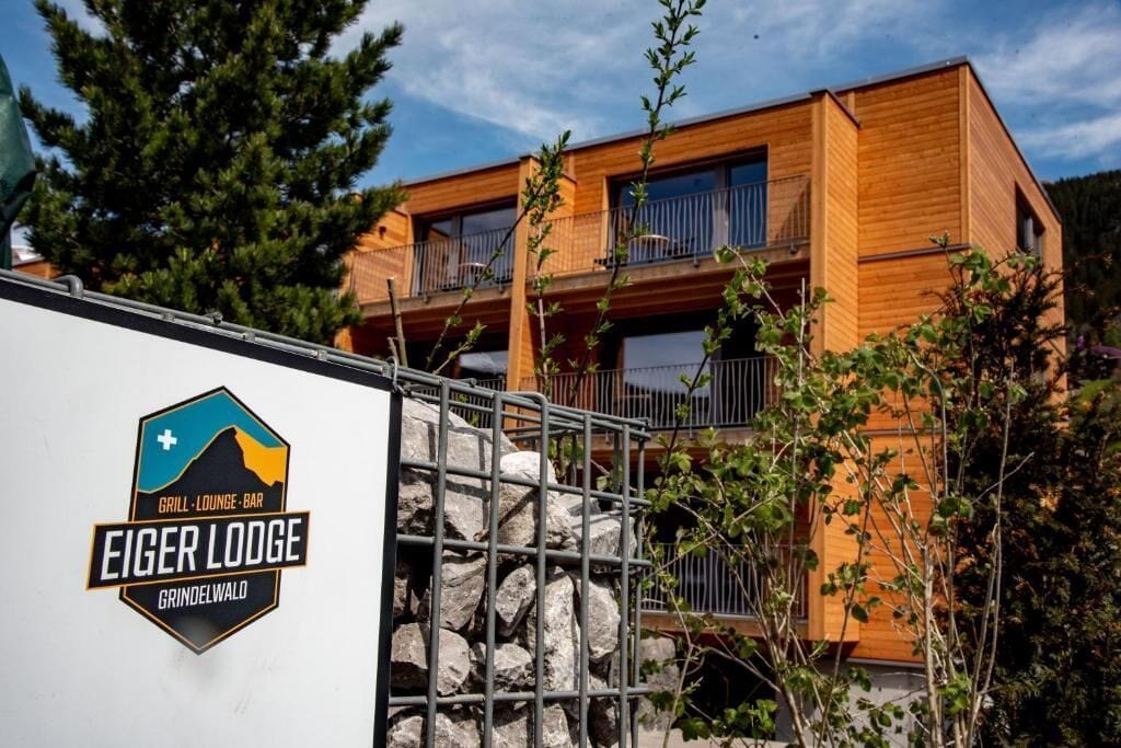 A two-story wooden building with balconies, a sign for 'Eiger Lodge Grindelwald,' and a metal fence.