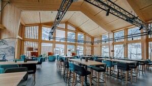 A spacious indoor area with high ceilings and large windows, featuring multiple tables and chairs arranged for dining or meetings. - Eiger Lodge Easy (Grindelwald)