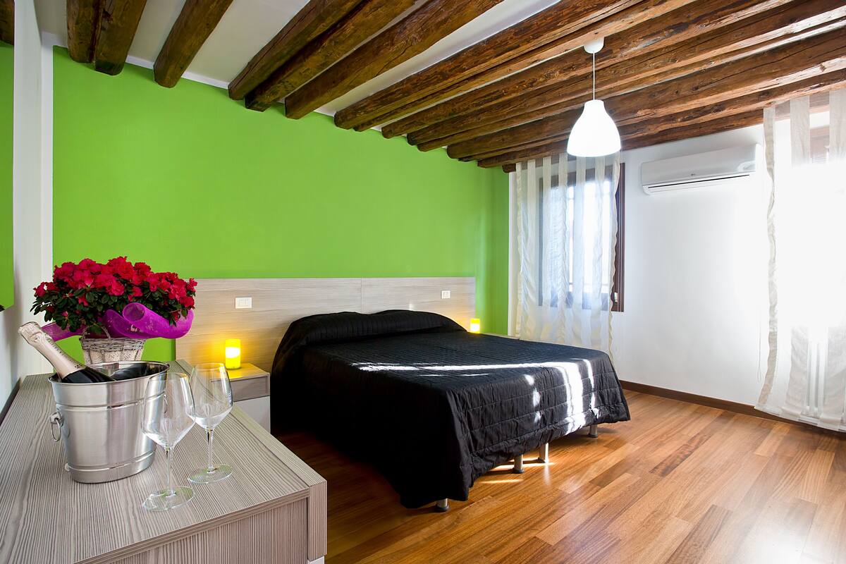 Una stanza con un letto, soffitto in legno, parete verde, finestra con tende e un tavolo con una bottiglia di champagne e bicchieri.