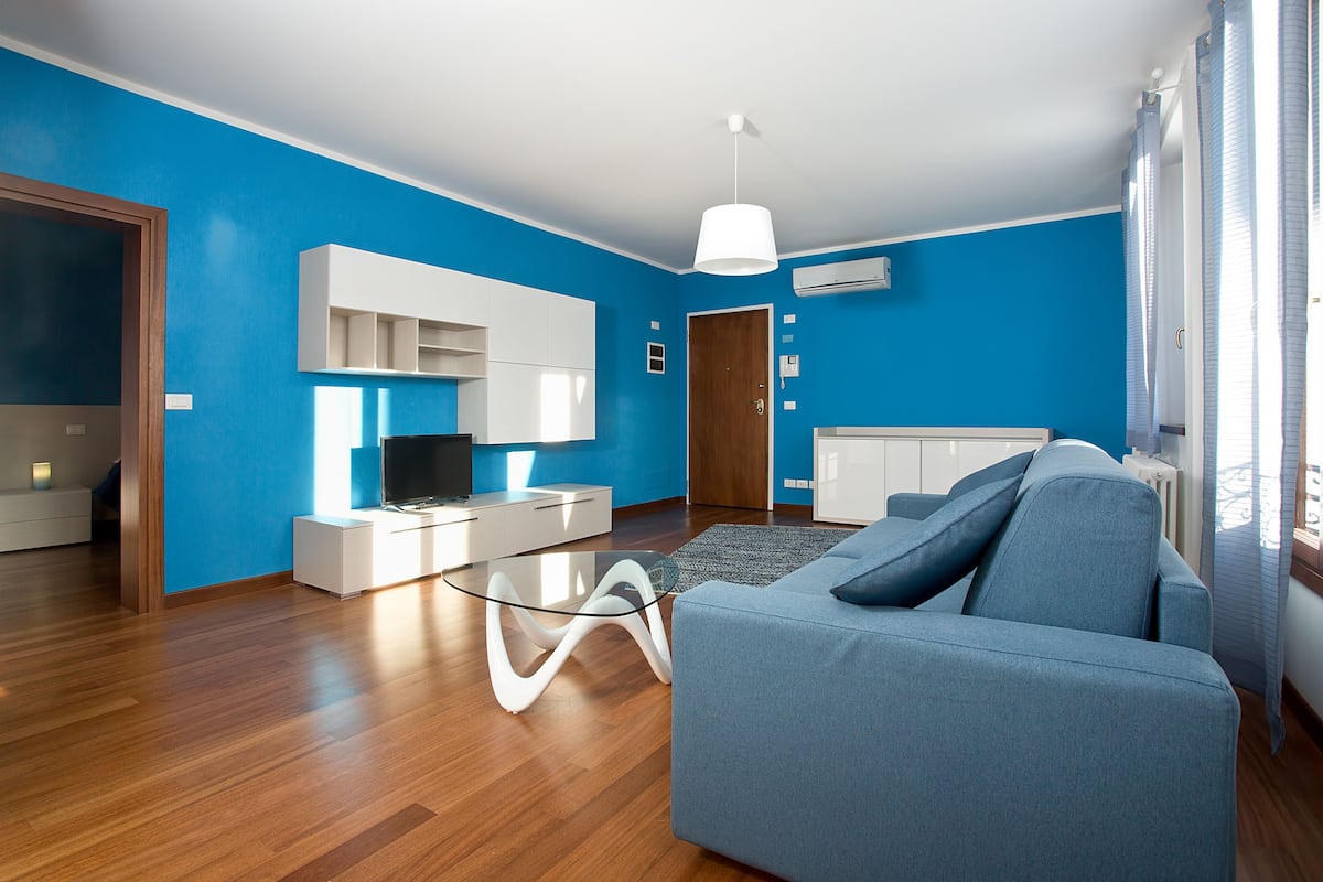 Un soggiorno moderno con parete blu, pavimento in legno, divano blu, tavolino da caffè in vetro, mobile TV bianco e televisore.