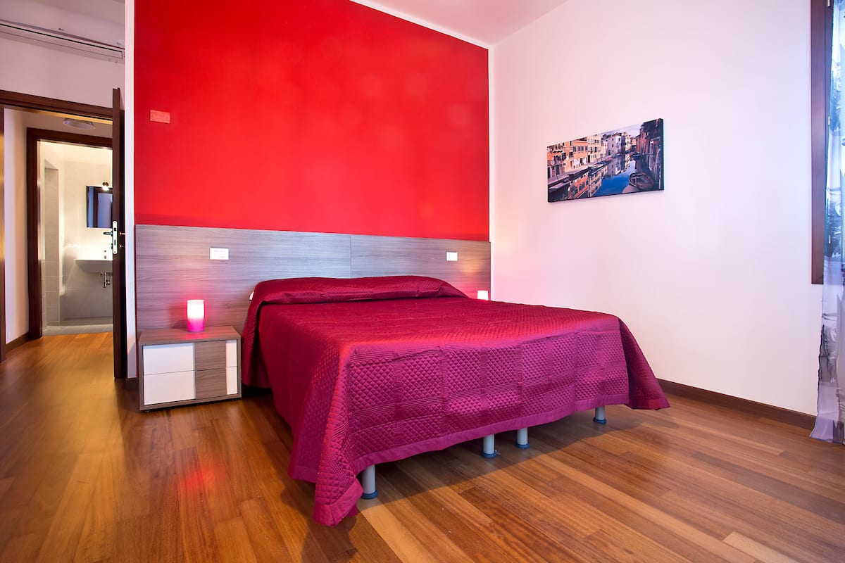 Una camera d'hotel moderna con una parete rossa a contrasto, un letto con coperta rosa, un comodino con lampada e un quadro raffigurante un panorama urbano.
