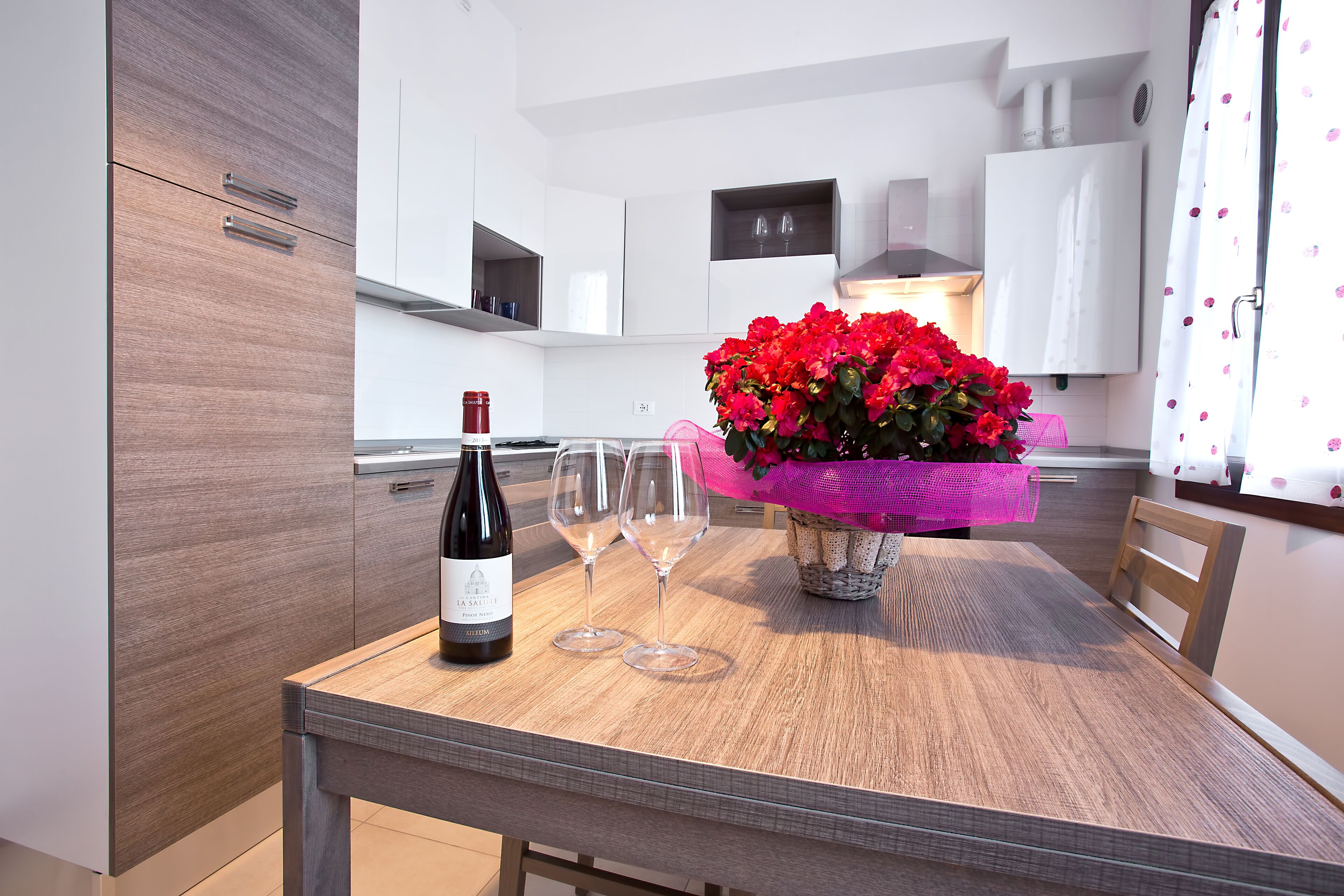 A modern kitchen with wooden cabinets, a dining table with two wine glasses and a bottle, and a vase with red flowers.
